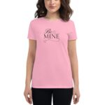 A woman wears a short-sleeved pink T-shirt featuring the text "Be MINE" in black script with small heart designs  standing against a neutral background.