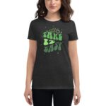 A dark shirt features the phrase "TAKE IT EASY" in playful green letters  surrounded by small stars and two cartoonish fruit icons  worn by a woman with shoulder-length hair.