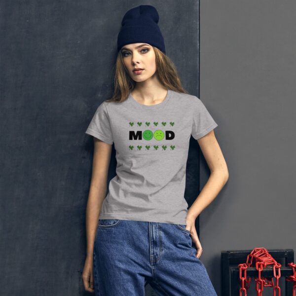 A woman stands confidently wearing a gray t-shirt with a "MOOD" graphic featuring two facial expressions and green leaves against a dark wall with red chains in the background.