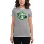 A woman wears a gray t-shirt featuring the word "EXPECTATION" surrounded by green shamrocks  standing against a neutral background. The design emphasizes a cheerful and vibrant theme.
