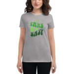 A woman is wearing a gray t-shirt with a large green graphic design that says "TAKE IT EASY" surrounded by small stars and abstract shapes  standing against a neutral background.
