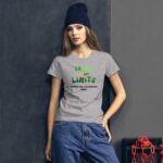 A young woman stands against a dark wall wearing a grey t-shirt with green and black text that reads DREAM with out LIMITS HAPPY ST PATRICKS 2024 paired with loose blue jeans and a navy beanie