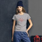 A young woman poses wearing a gray t-shirt with the text "My 1st Valentine's Day" against a dark textured wall  with a beanie on her head and denim jeans.