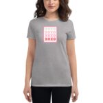 A woman is wearing a gray t-shirt featuring a colorful design of the word "XOXO" repeated in a square pattern on the front while standing against a neutral background.