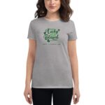 A woman wears a gray t-shirt displaying green text that reads not lucky just blessed and happy St. Patrick's Day surrounded by small shamrock illustrations in a neutral background.
