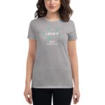 A woman wears a gray t-shirt featuring green and white text that reads LUCKY HAPPY ST. PATRICK'S DAY surrounded by decorative shamrocks and stars in a neutral background
