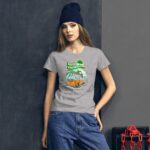 A woman wearing a grey t-shirt with a colorful graphic of a vintage van and palm trees stands against a dark wall she is also wearing a beanie and denim jeans.