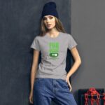 A young woman stands against a dark wall wearing a gray t-shirt with green text that reads FUN MODE ON HAPPY ST PATRICK'S DAY paired with denim shorts and a dark beanie.