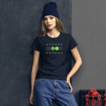 A young woman stands against a dark gray wall wearing a black t-shirt with "MOOD" and green emojis surrounded by small green decorations paired with loose blue jeans and a blue beanie.