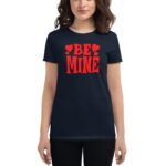 A woman stands wearing a navy t-shirt featuring bold red text that reads BE MINE with heart shapes on either side suggesting a romantic theme in casual attire.