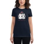 A woman wears a dark t-shirt featuring two cartoon cats expressing affection with hearts above them and the text Happy Valentine's Day below the design in a casual setting.