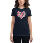 A woman wearing a dark t-shirt with a floral heart design stands against a neutral background showcasing vibrant flowers including pink roses and green leaves in a casual pose.