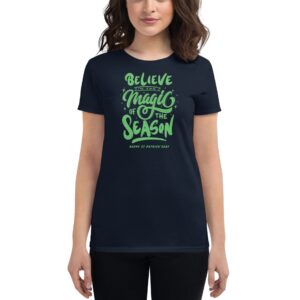 A woman is wearing a dark t-shirt featuring green text that reads Believe in the Magic of the Season Happy St. Patrick's Day creating a festive holiday-themed appearance.