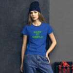 A young woman stands against a dark wall wearing a blue t-shirt with the text KEEP IT SIMPLE in green letters while sporting a blue beanie and light blue jeans.