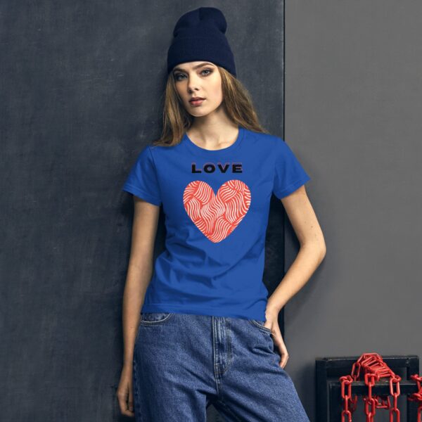 A young woman stands against a textured grey wall wearing a bright blue shirt featuring a red heart with fingerprint designs and the word LOVE above it. She has long hair and a dark beanie.