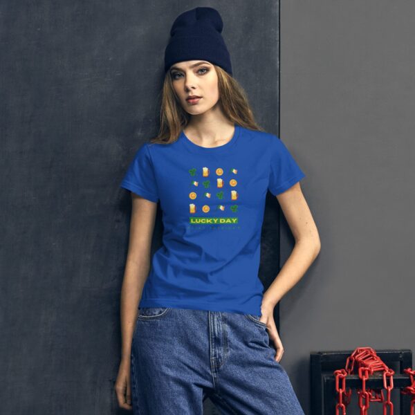 A woman wears a blue t-shirt featuring a colorful graphic with the text LUCKY DAY. She stands against a dark backdrop  complemented by casual jeans and a navy beanie.