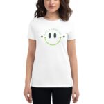 A woman wears a white t-shirt featuring a green smiley face graphic and text that says HAVE A LUCKY DAY surrounded by small shamrocks  standing against a plain background.
