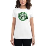 A woman wears a white t-shirt featuring the word "EXPECTATION" in green with a leafy design around it  standing against a neutral background.