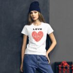 A young woman stands against a dark wall wearing a white t-shirt with a red heart graphic and the word LOVE above it while dressed in casual blue jeans and a black beanie.