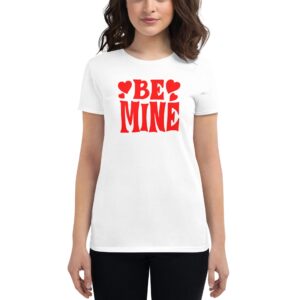 A woman stands wearing a white t-shirt emblazoned with red text that reads BE MINE surrounded by small red heart shapes in a neutral background.