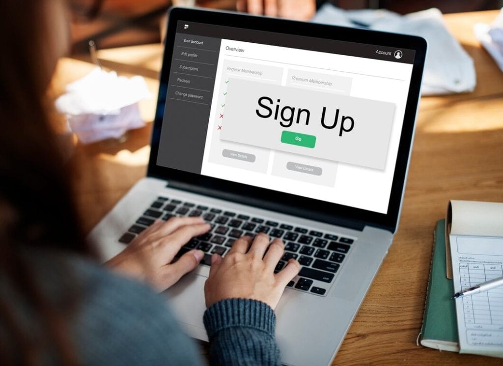 A laptop displays a sign-up screen while a person types on the keyboard in a cozy indoor setting with a wooden table and a notebook nearby.
