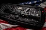 A black tactical rifle is resting inside a zippered carrying case with padded inserts while positioned on a folded American flag displaying red white and blue colors with white stars.