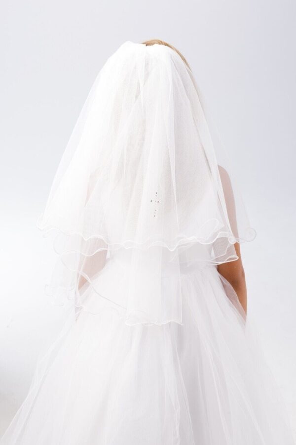 A child wears a flowing white dress and a sheer veil  standing still against a plain light background  suggesting a moment of innocence and celebration.
