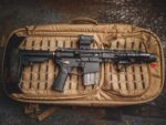 A black firearm rests horizontally atop a tan padded bag with a grid pattern The firearm features a scope and flashlight positioned on top with a textured grip and a magazine attached