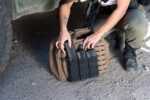 A person is kneeling by a brown tactical backpack securing firearm magazines into elastic pouches positioned on the bag's side the surroundings are rocky and dusty under a vehicle