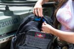 A person is opening a black backpack to retrieve sunglasses in a bright outdoor setting near military equipment. The backpack features a patch labeled "ADVANCED WARRIOR SOLUTIONS."