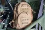 A tan backpack rests on a vehicle seat next to a steering wheel indicating it's ready for use in an outdoor or tactical context with visible gear attachment points.