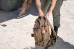 A person with long hair is opening a brown backpack on a gravel surface while standing near a large vehicle  revealing a water bottle in the side pocket.
