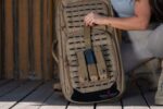 A tan tactical backpack with a structured front is being adjusted by a person who reaches for a device secured in a pocket while crouching on a wooden surface.