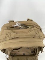 A tan tactical backpack is open with a clear plastic object partially visible inside its top compartment while resting on a flat surface with a neutral background.