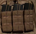 Three black magazines are secured within tan fabric pouches on a tactical vest background showcasing a grid pattern indicating military or outdoor equipment in a low-light setting.