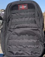 A black backpack with multiple straps and pockets lies flat against a surface showcasing a smooth fabric texture and a logo patch labeled Advance Warrior Solutions in a bright red rectangular border.