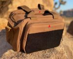 A tan tactical bag with multiple compartments sits on a rocky surface in a desert setting reflecting sunlight with a black fabric section on the front and a logo in red.