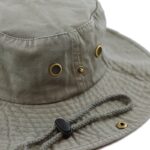 A wide-brimmed olive green bucket hat features eyelet vents and a braided chin strap with a toggle for adjustment resting on a textured surface.