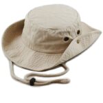 A beige bucket hat rests on a flat surface featuring a round crown and wide brim with vent holes the adjustable chin strap is attached and hangs loosely along the side
