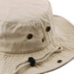 A beige bucket hat rests on a flat surface featuring ventilation eyelets and an adjustable chin strap indicating it is designed for outdoor use and sun protection.