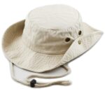 A beige bucket hat lies flat with a wide brim and rounded top featuring eyelets. It has an adjustable chin strap for securing during outdoor activities.