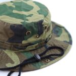 A camouflage bucket hat rests with its wide brim facing outward showcasing a mix of green and brown patterns while a drawstring lies loosely attached around the base