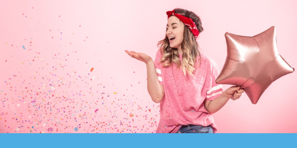 A woman wearing a red paisley bandana smiles while holding a metallic star-shaped balloon. She blows confetti into the air, set against a pink background.