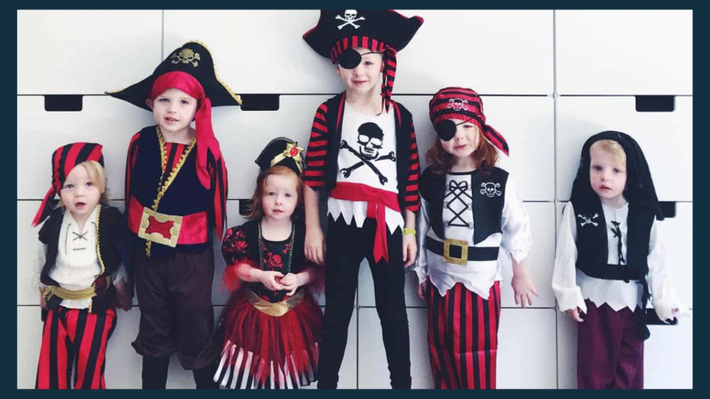 Six children wearing colorful pirate costumes and vibrant bandanas stand in a row, smiling and playful. Their outfits add charm and energy to the cheerful scene, set against the simple backdrop of a white storage unit.