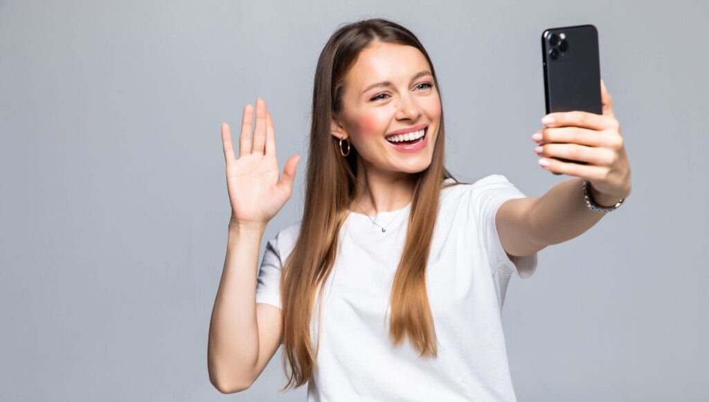 A smiling woman in a white t-shirt takes a selfie with a smartphone, waving with her other hand. The background is a neutral gray, conveying a joyful mood.