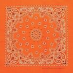 An orange bandana features intricate white paisley designs and floral motifs arranged symmetrically over the entire fabric creating a vibrant decorative pattern suitable for various uses.