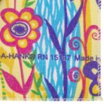 A colorful fabric features vibrant flowers and swirls against a yellow background. Text reads "A-HANK® RN 1587 Made in." The design exudes a playful  cheerful aesthetic.