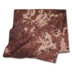 A piece of fabric with a brown  mottled pattern lies flat with some folds creating texture and depth in a neutral setting  suggesting it could be used for clothing or decoration.