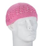 A pink crochet cap sits atop a smooth white mannequin head showcasing an intricate pattern with holes throughout creating a lightweight appearance in a neutral backdrop.