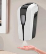 A wall-mounted soap dispenser releases liquid soap into an extended hand positioned below it in a clean indoor environment with a light-colored wall and a window in the background.